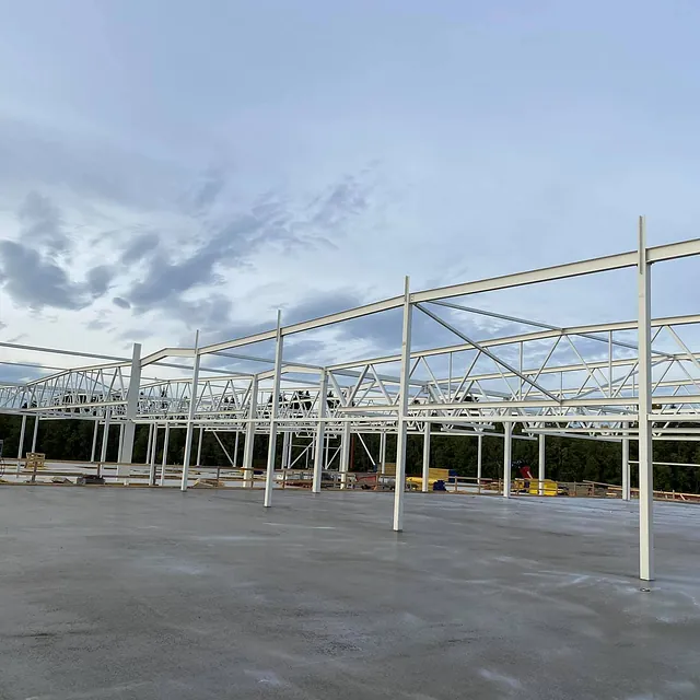 Stålramme til bygg under oppføring på betongdekke, med trær og overskyet himmel rundt.